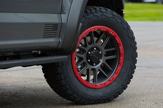 Close-up of a rugged off-road tire with distinctive red rim on a sleek ROUSH Raptor, parked on a paved surface. Its high-performance suspension hints at its readiness for exhilarating off-road racing adventures.