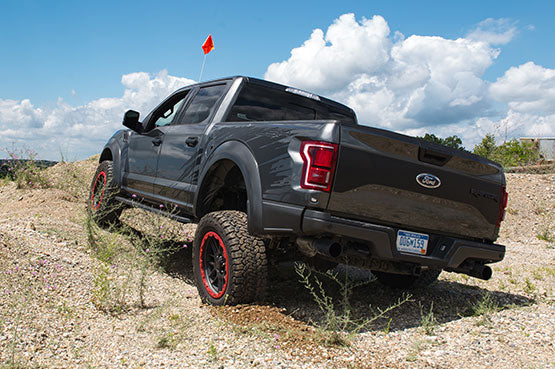 A black ROUSH Raptor pickup truck powers uphill on a rocky terrain with off-road suspension, beneath a cloudy blue sky, sporting Bead Protection Wheels and a red flag fluttering at the back.
