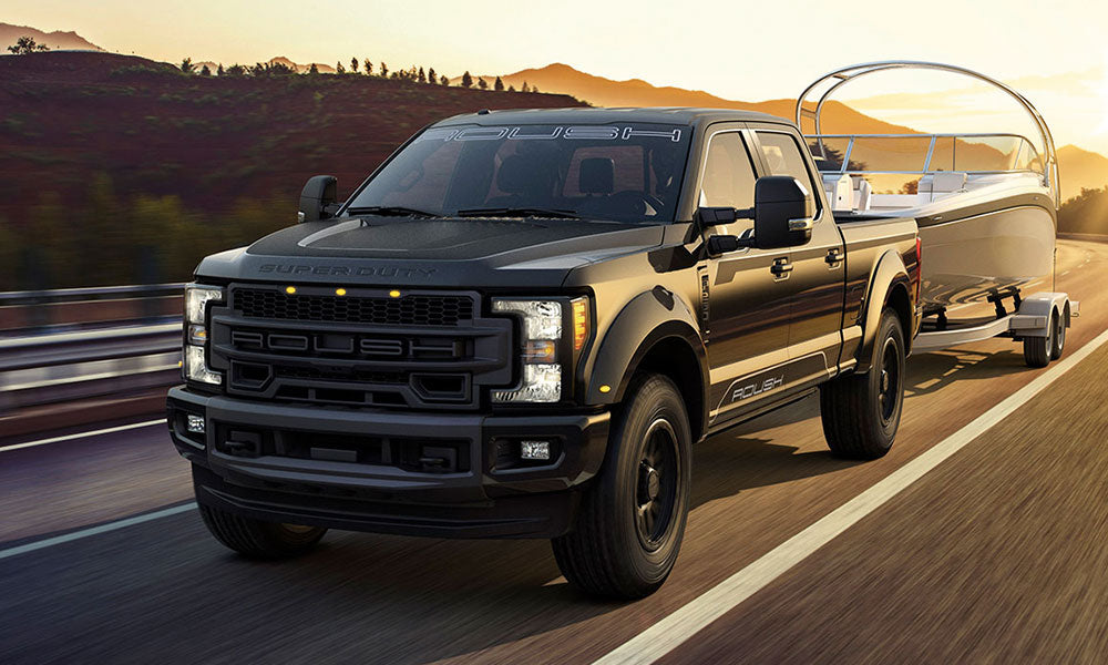 A black ROUSH Super Duty pickup truck, powered by a Turbo Diesel V8, gracefully tows a boat on the highway at sunset, with hills forming a perfect backdrop.