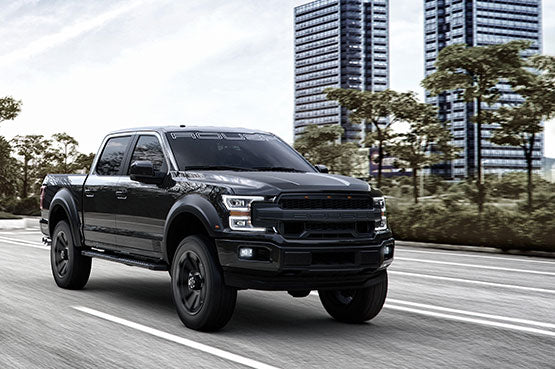 A black ROUSH F-150 pickup truck with impressive performance upgrades drives on a road, with blurred city buildings and trees in the background.