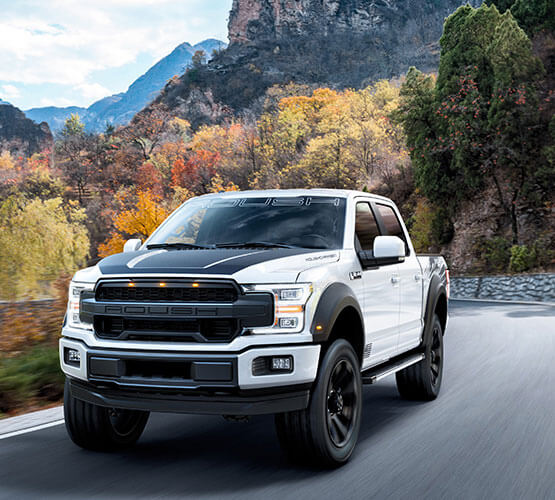 A ROUSH F-150 SC with a performance exhaust system roars down a scenic mountain road, surrounded by vibrant autumn foliage.