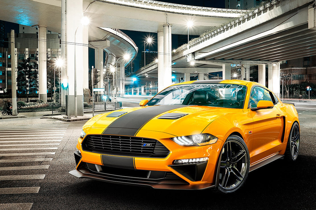 A bright yellow Mustang with black racing stripes and a roaring 5.0L V8 engine is parked on a city street beneath elevated highways at night.