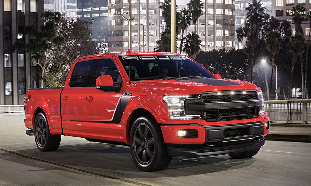 A red pickup truck, equipped with a supercharged 650-horsepower V8, cruises down a city street at night, with buildings and palm trees silhouetted in the background.