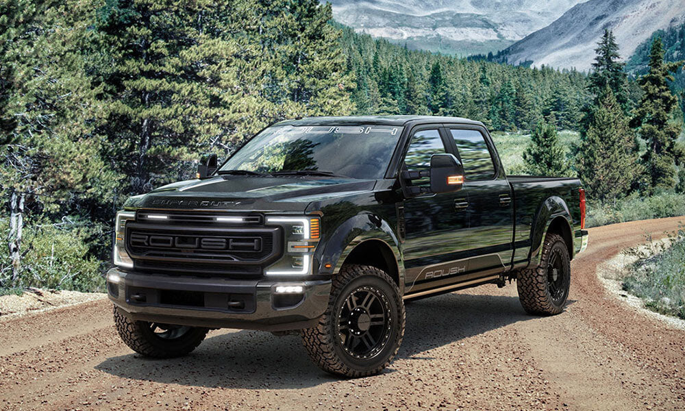 A black pickup truck equipped with all-terrain tires on a dirt road is surrounded by a dense forest and majestic mountains in the background.