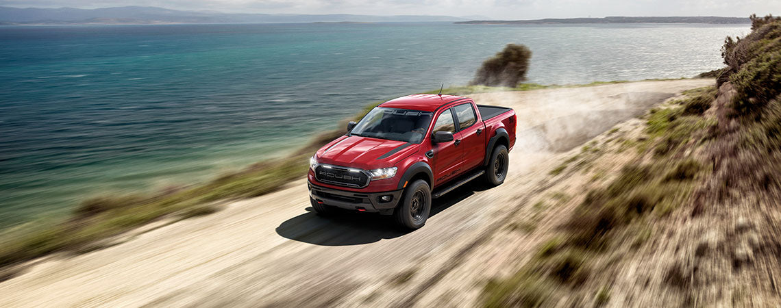A ROUSH Ranger, boasting off-road prowess and a high-performance suspension system, drives along a coastal dirt road with the ocean as its stunning backdrop.