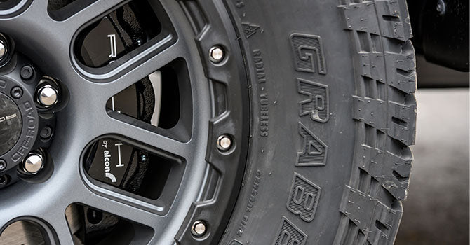 Close-up of a rugged tire with the word "GRABBER" imprinted on the sidewall, mounted on a black alloy wheel, highlighting its off-road capable design perfect for a performance truck.