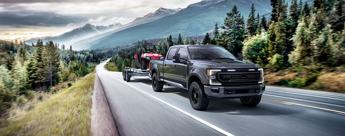 A dark gray ROUSH Super Duty pickup truck, equipped with all-terrain tires, pulls a trailer carrying an ATV on a scenic mountain road, surrounded by trees and distant peaks.