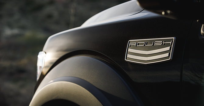 Close-up of the front side of a black ROUSH F-150 vehicle featuring a badge with the word "Roush," a three-stripe design, and hints of off-road performance enhancements.