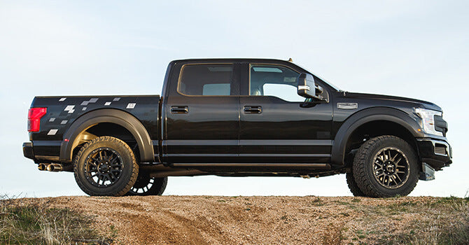 Side view of a black ROUSH F-150 pickup truck with rugged tires parked on a dirt mound under a clear sky, showcasing its off-road performance.