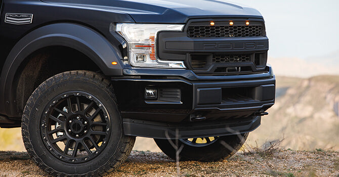 Close-up of a black ROUSH F-150 pickup truck's front section and wheel, featuring a prominent grille and rugged terrain tires, parked on a dirt surface with a blurred natural background. This off-road performance beast is ready to tackle any adventure.