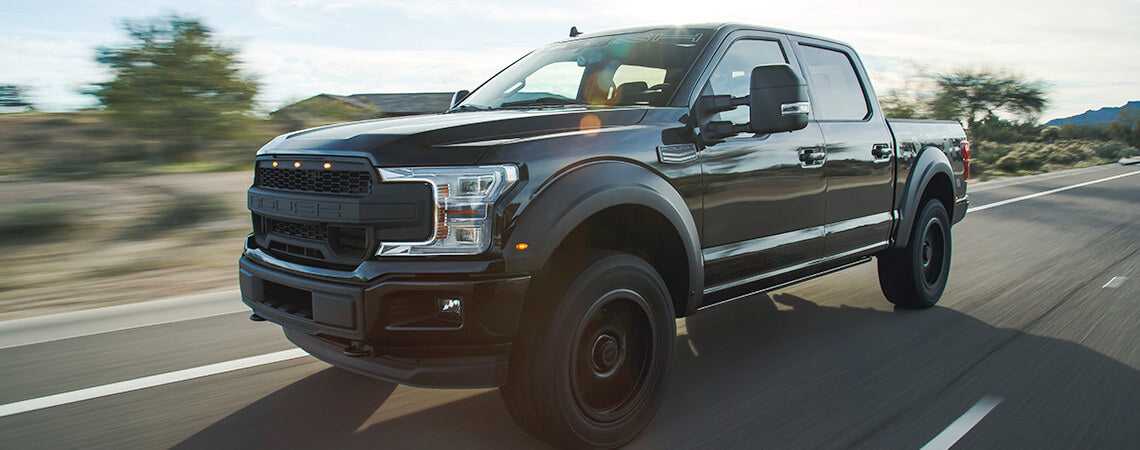 A black ROUSH F-150 pickup truck drives on a highway, showcasing its off-road performance capabilities against a desert landscape under a clear sky.