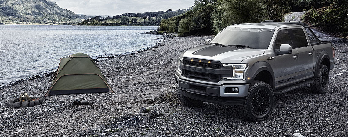 A gray ROUSH F-150 pickup truck, known for its off-road capability, is parked on a rocky lakeshore next to a green tent and a small campfire. Mountains and trees provide a stunning backdrop.