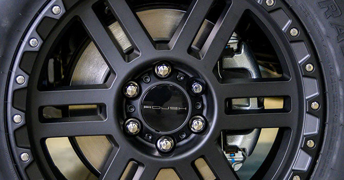 Close-up of a black alloy wheel with a visible braking system behind it. The center hub, adorned with bolts and the ROUSH F-150 logo, hints at its off-road capability.