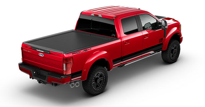 A red pickup truck, featuring ROUSH Super Duty enhancements, showcases a covered bed, black accents, and large wheels with an advanced performance suspension system. Its robust design is complemented by all-terrain tires, captured from the rear three-quarter angle.