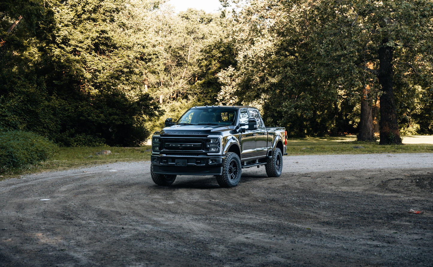 A black Roush Super Duty pickup truck with a performance suspension is parked on a dirt road surrounded by trees and greenery on a sunny day.