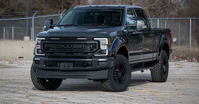 A black pickup truck with "ROUSH" on the grille stands proudly on a paved surface, featuring 20-inch ROUSH wheels. In the background, trees and a chain-link fence complete the scene.