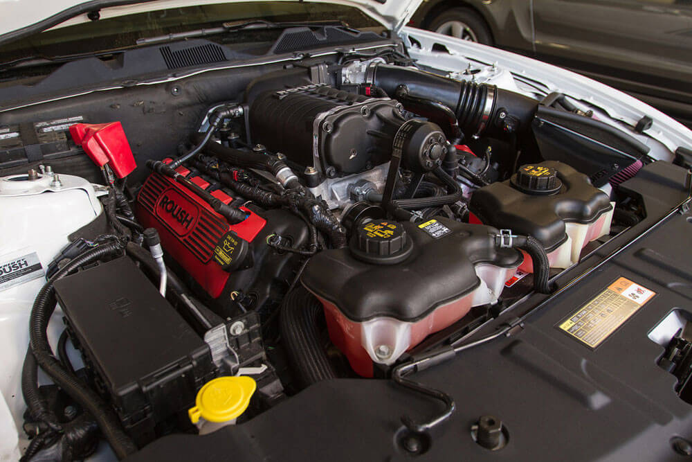 Close-up of a car engine compartment featuring a Roush Mustang Supercharger, showcasing Eaton's TVS Technology with red components labeled "Roush" and several black fluid reservoirs.