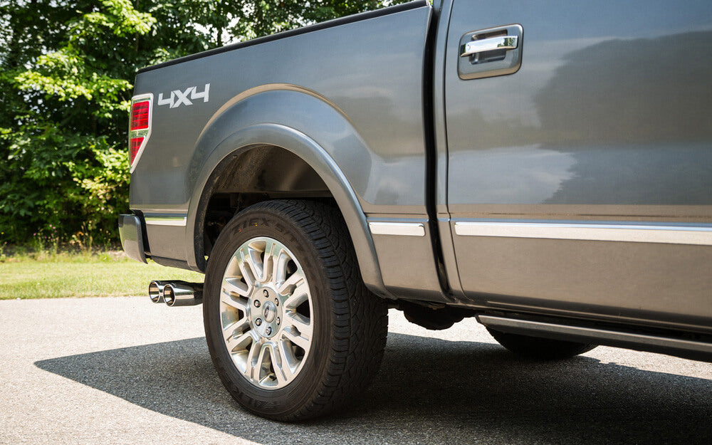 Close-up of a gray 4x4 truck's rear wheel and side, parked on a paved road with trees in the background. The gleam of a stainless steel exhaust system catches the eye, adding a sleek touch to the rugged vehicle.