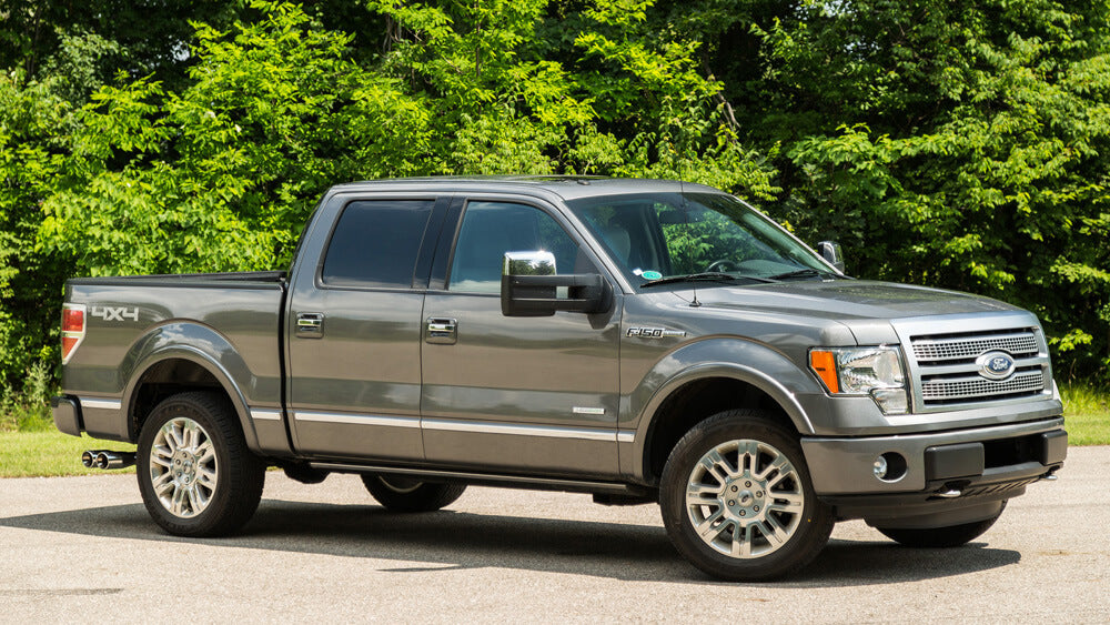 A gray Ford F-150 pickup truck, enhanced with a sleek Roush exhaust, is parked on a road with lush green trees in the background.