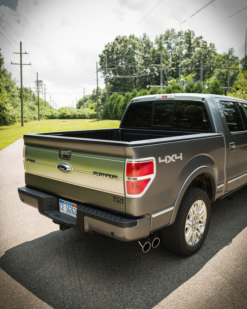 A silver Ford F-150 Platinum truck glides along the road, its robust stainless steel Roush exhaust system quietly humming amidst a backdrop of trees and power lines.