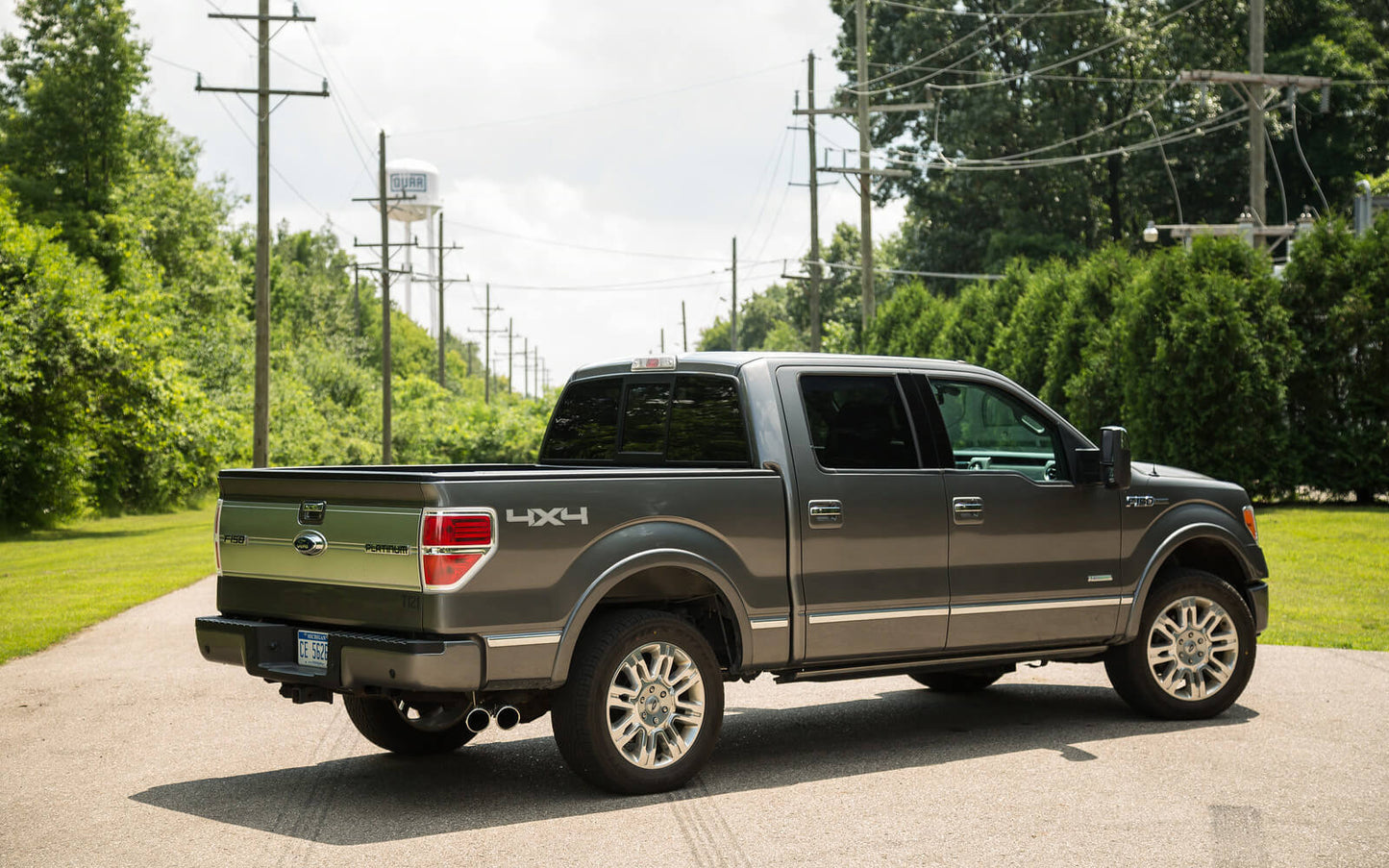 A gray pickup truck with a gleaming stainless steel F-150 exhaust system is parked on a paved road, surrounded by lush greenery and power lines.