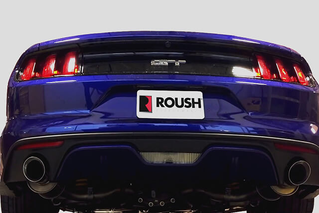 Rear view of a blue sports car with dual exhausts, illuminated tail lights, and a license plate displaying the Roush logo, featuring the aggressive sound of a Roush Axle Back Exhaust for 2015-2017 Mustang models.