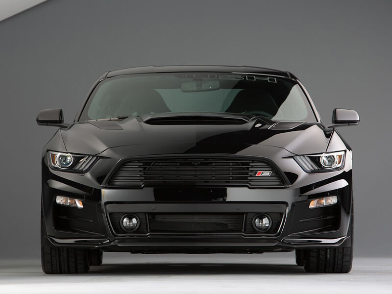 A black Ford Mustang with a sleek design, featuring a Roush Mustang Front Fascia Kit from Roush Performance Products, Inc. and aggressive headlights, parked in a studio setting.