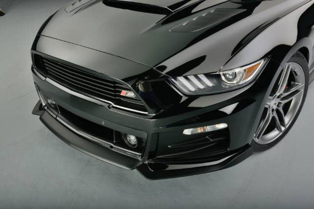 
                  
                    Close-up of a black sports car's front end, featuring sleek headlights and a Roush Performance Products, Inc. 2015-2017 Roush Mustang Complete Front Fascia Kit - Raw Unpainted, on a gray floor.
                  
                