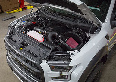 A pickup truck with its hood open, revealing the engine and various components in a garage or workshop setting, proudly showcasing a high-performance cold air intake installed for boosted efficiency.
