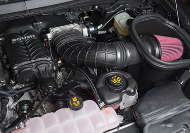 A car engine equipped with the Roush F-150 Phase 2 Upgrade Kit is displayed, showcasing a powerful 650 HP Roush supercharger, complemented by a pink air filter and featuring coolant and oil reservoirs.