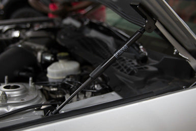 Close-up view of a car hood propped open by Roush hood struts, revealing parts of the engine underneath. Perfect for Mustang models 2015-2023, these gas-charged struts offer a sleek and reliable alternative to traditional metal rods.