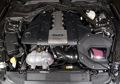 View of a Mustang 5.0L engine with a sleek black cover labeled "5.0," showcasing various components, including a striking red air filter that's part of the Cold Air Kit.