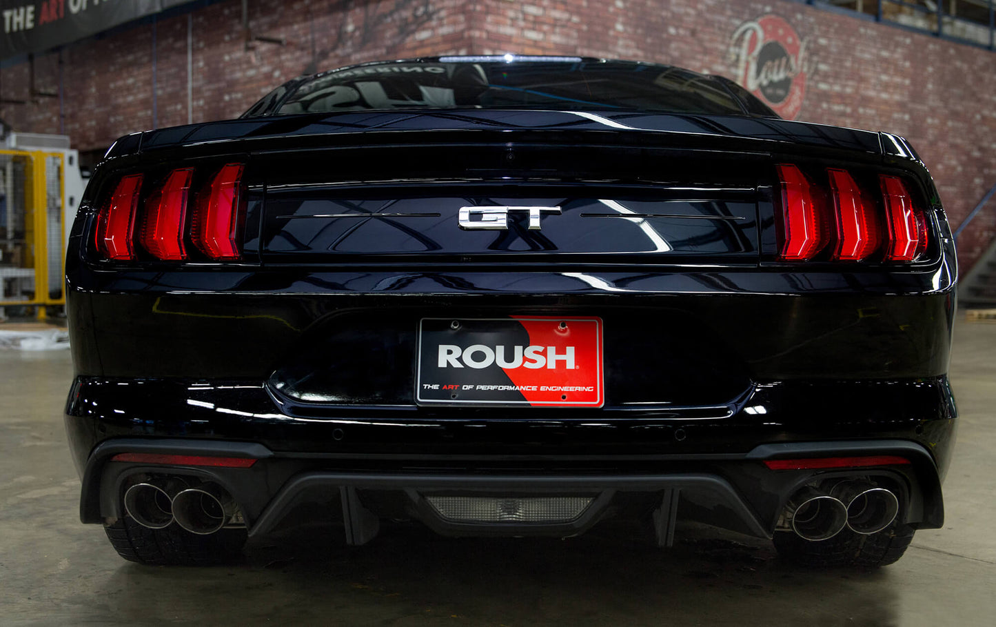 Rear view of a black 2018-2023 Mustang GT sporting a "GT" badge and a "Roush" branded license plate, complemented by the sleek T-304 stainless steel Roush Performance Axle-Back Exhaust System under a garage setting.