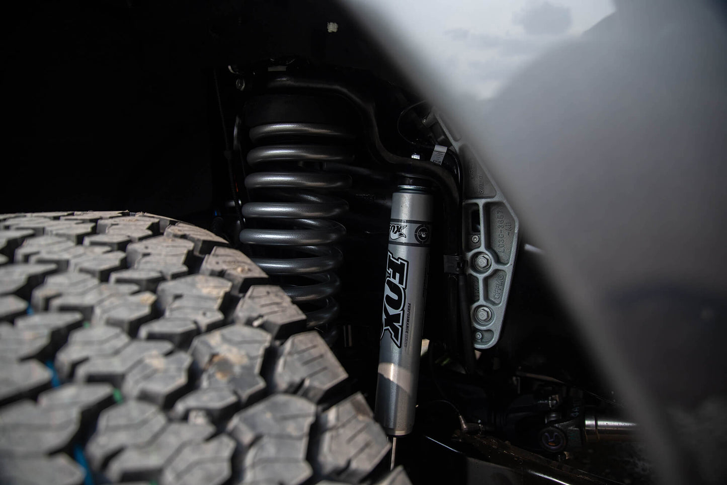 Close-up of a vehicle's suspension system featuring a coil spring, Fox shocks from the Roush Suspension Kit, and part of an off-road tire with a rugged tread pattern.