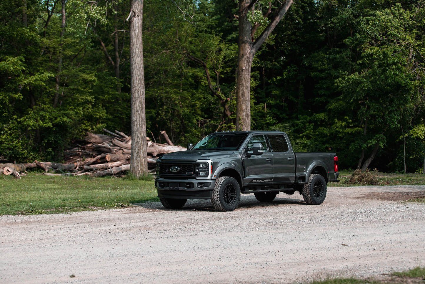 A black Ford Super Duty pickup truck is parked on a dirt road with a forested area and a pile of logs in the background, featuring advanced Fox shocks for superior off-road handling.