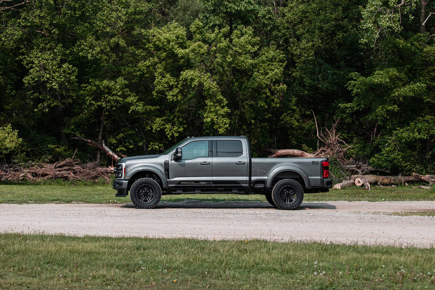 A gray Ford Super Duty pickup truck with a crew cab is parked on a gravel road in front of dense, green woodland, equipped with Roush Suspension Kit for enhanced performance.