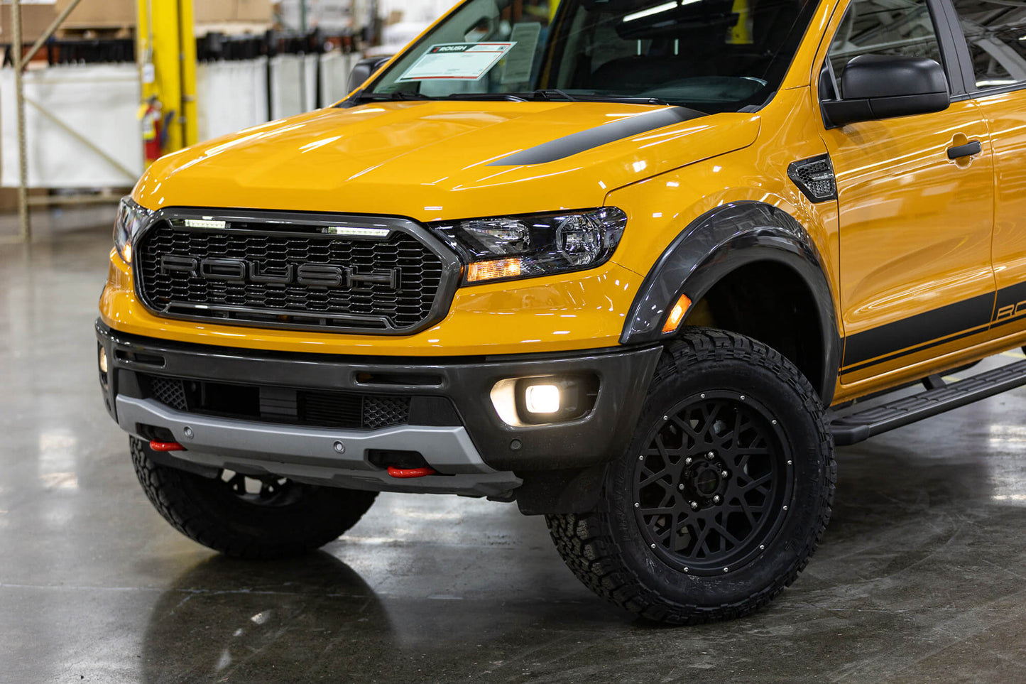 Close-up of a yellow 2022 Roush Ranger's front end, showing the grille, headlight, and part of the tire in an indoor setting. This Roush Performance vehicle showcases its impressive off-road capability even at first glance.