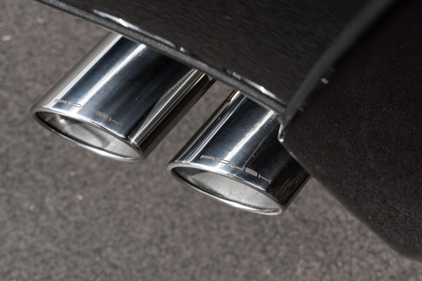 Close-up view of dual chrome exhaust tips on a black vehicle, marked with the Porsche logo, set against a gray pavement background, adjacent to the ROUSH High-Flow Front Grille.