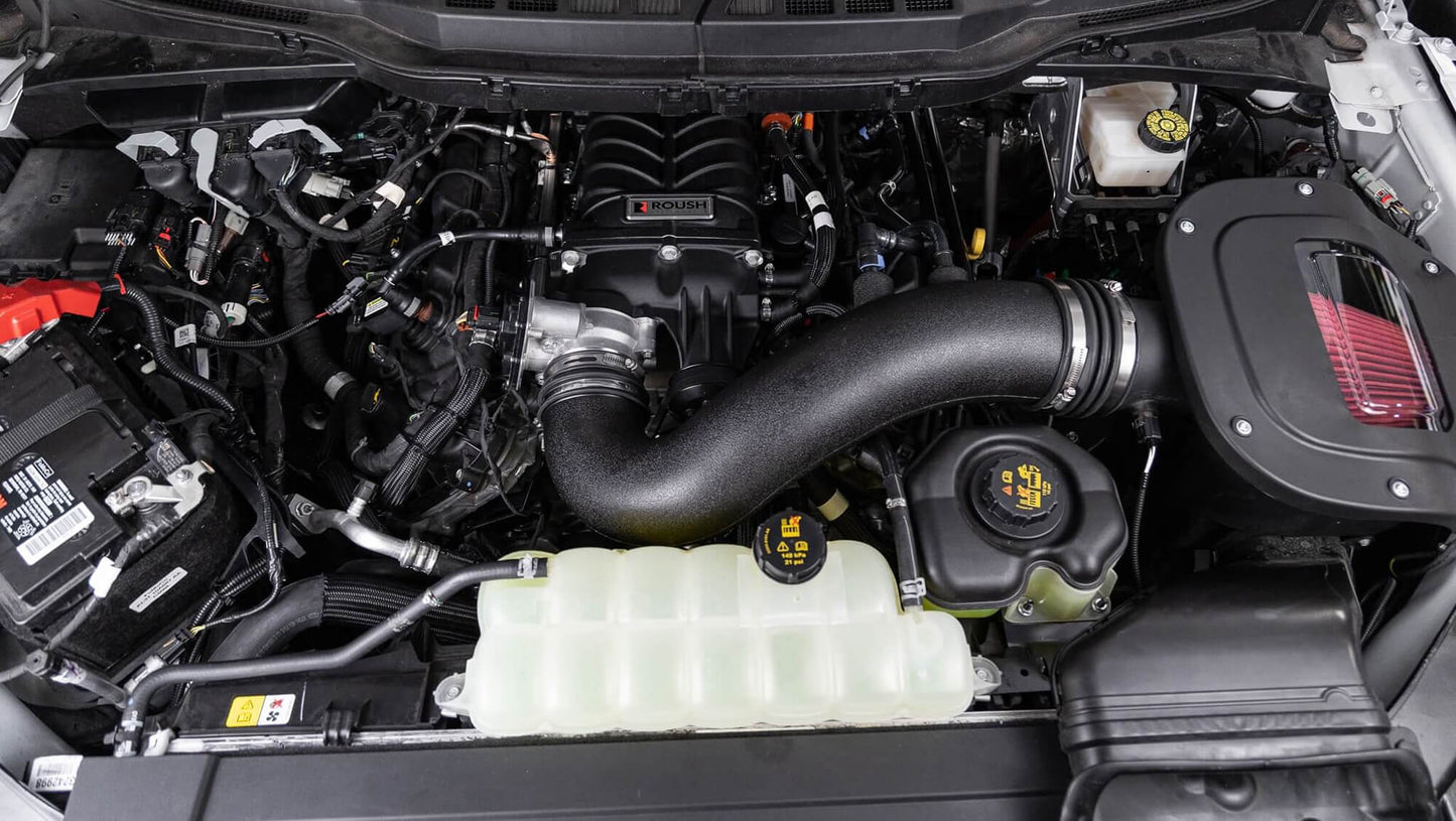 A close-up view of a car engine compartment showcasing various components, including the Roush Performance F-150 Supercharger Kit, air intake, battery, coolant reservoir, and other engine parts that contribute to its impressive 705 horsepower.