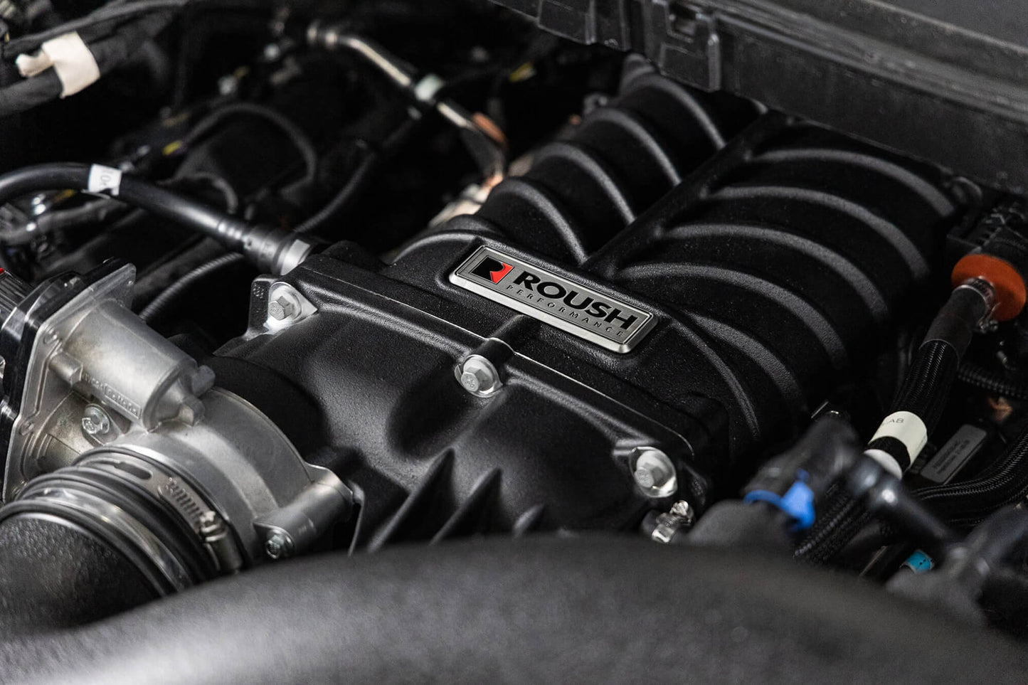 Close-up of a Roush Performance supercharger installed in a car engine bay. The supercharger, part of the F-150 Supercharger Kit, is prominently labeled and surrounded by various engine components and wiring. This powerhouse boosts the engine to an impressive 705 horsepower.