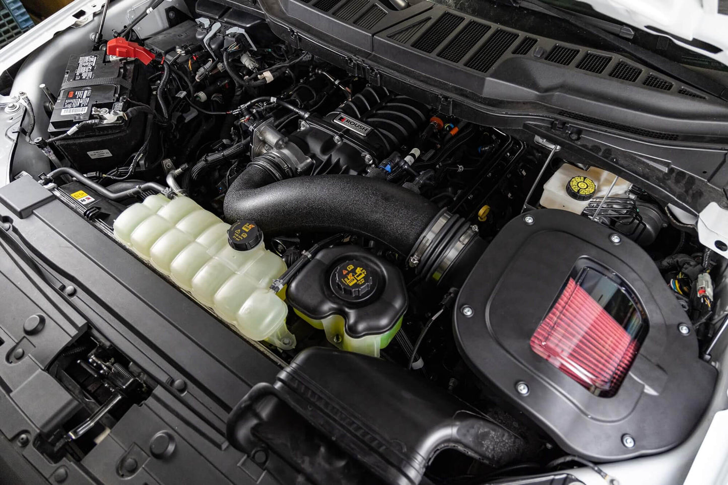 A close-up view of a car engine bay showcasing various components, including the air intake system, battery, and coolant reservoir spotlighting the impressive Roush Performance F-150 Supercharger Kit that delivers an incredible 705 horsepower.
