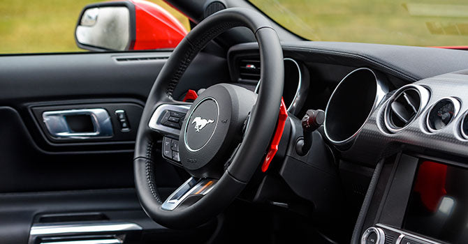 View of a Mustang's interior showcasing a black steering wheel with the iconic running horse logo, dashboard, and air vents. Red accents are visible on the paddle shifters and car door, highlighting its ROUSH performance heritage.