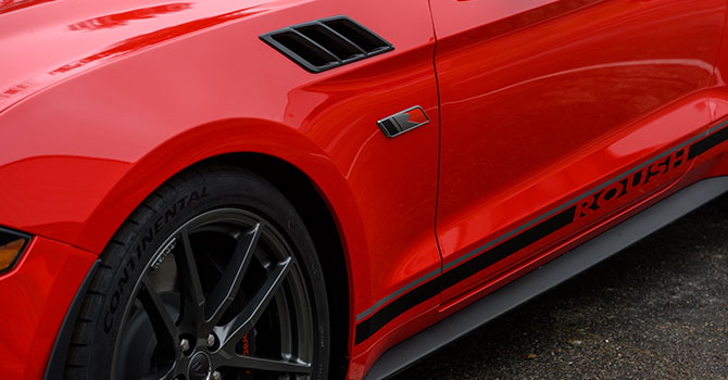 Close-up of the side of a red ROUSH Mustang sports car with black lettering and sleek black wheels glistening on a wet road, embodying sheer performance.
