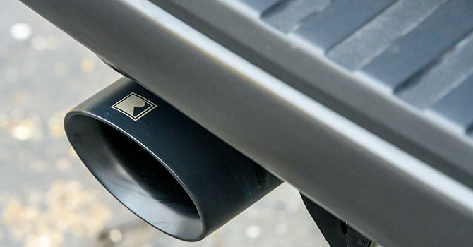 A close-up of a black exhaust pipe with a logo positioned beneath the dark bumper of an off-road capable vehicle, embodying the rugged spirit of a ROUSH Raptor.