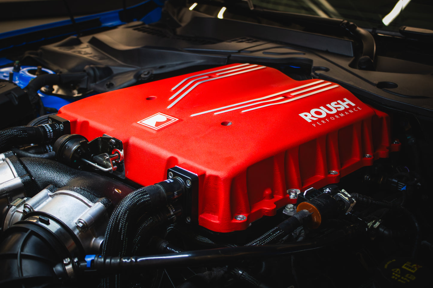 Close-up of a car engine with a red cover labeled "ROUSH Performance," showcasing the impressive power of an 810-horsepower Roush Mustang Supercharger. The intricate network of cables and components work harmoniously with the TVS R2650 Supercharger for peak performance.