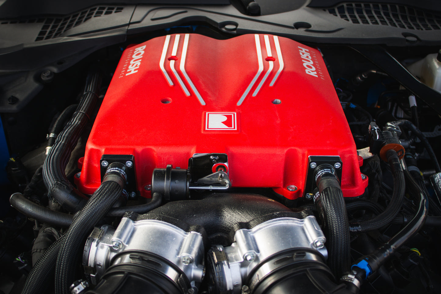 Close-up of a red Roush S650 Mustang engine cover with visible hoses and components under a car hood, showcasing its impressive 740 horsepower Supercharger Kit.