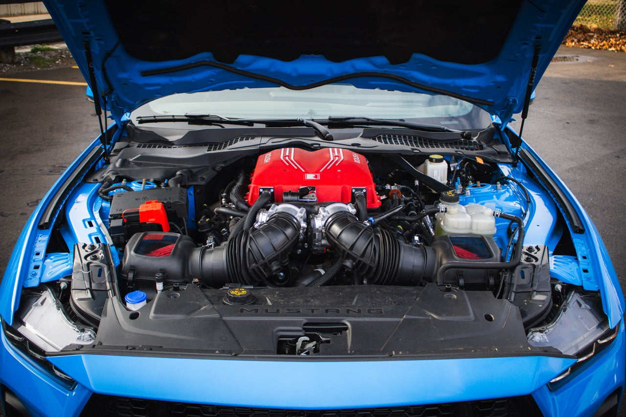 The open hood of a blue Roush S650 Mustang reveals a powerful red engine with dual air intakes, equipped with a supercharger kit that boosts it to an impressive 740 horsepower.