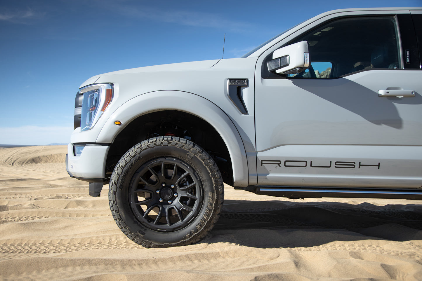 A light gray pickup truck with ROUSH branding and aggressive styling is parked on sandy terrain, under a clear blue sky, its 20-inch Matte Black Roush F-150 wheels completing the bold look.