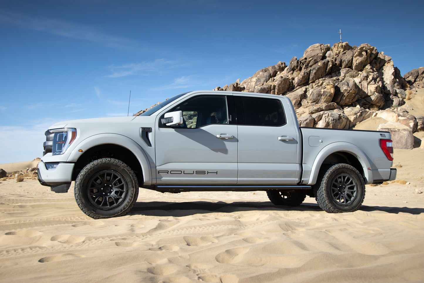 A white pickup truck with aggressive Roush styling and "Roush" lettering parked on sandy terrain, its 20-inch wheels glinting under the clear blue sky, rocky hills standing robust in the background.