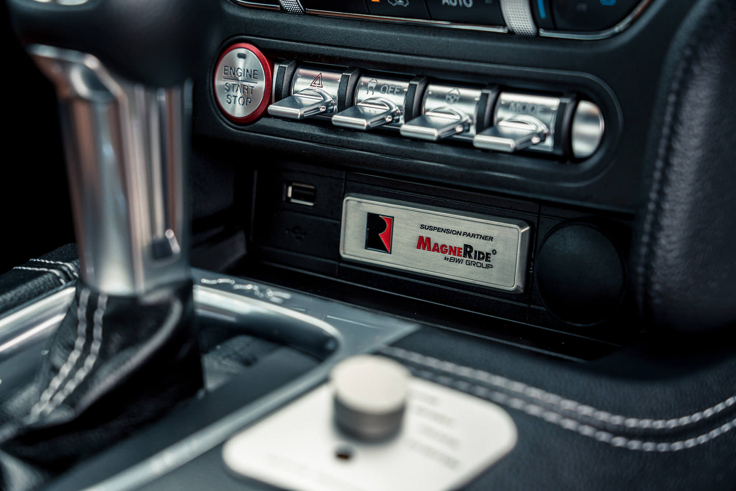Close-up of a Mustang's center console featuring a gear shift, engine start/stop button, toggle switches, and a suspension system with "MagneRide" for enhanced performance.